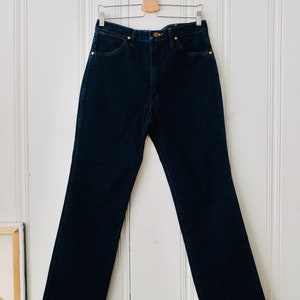 Peut inclure: Un jean en denim bleu fonc&eacute; suspendu &agrave; un cintre en bois contre un mur &agrave; panneaux blancs. Le jean a une conception classique &agrave; cinq poches avec une fermeture &agrave; bouton et &agrave; glissi&egrave;re. Le jean est de couleur indigo fonc&eacute;.