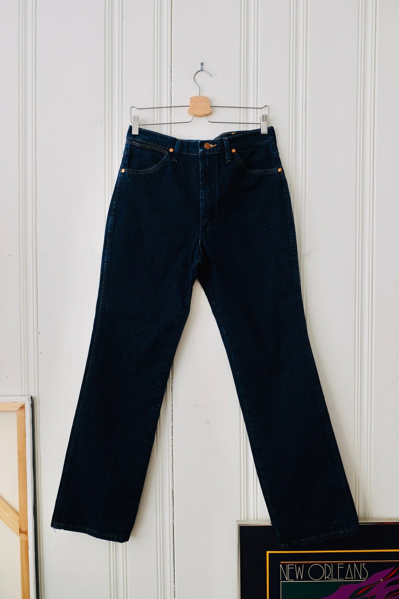 Peut inclure: Un jean en denim bleu fonc&eacute; suspendu &agrave; un cintre en bois contre un mur &agrave; panneaux blancs. Le jean a une conception classique &agrave; cinq poches avec une fermeture &agrave; bouton et &agrave; glissi&egrave;re. Le jean est de couleur indigo fonc&eacute;.
