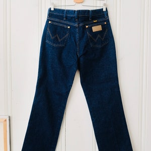 Peut inclure: Un jean en denim bleu fonc&eacute; suspendu &agrave; un cintre en bois. Le jean pr&eacute;sente le logo Wrangler sur la poche arri&egrave;re et une conception classique &agrave; cinq poches. Le jean est de style droit.