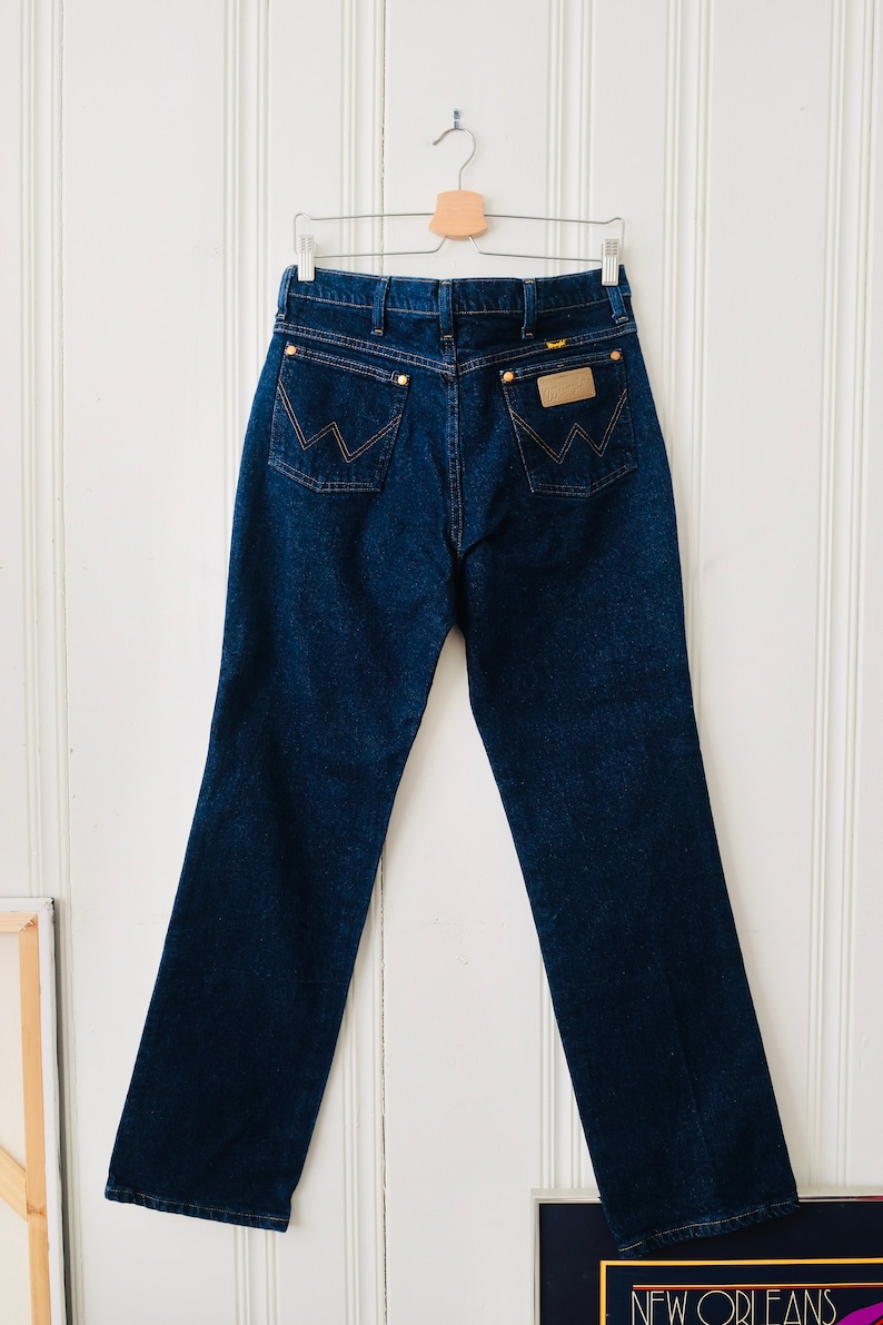 Peut inclure: Un jean en denim bleu fonc&eacute; suspendu &agrave; un cintre en bois. Le jean pr&eacute;sente le logo Wrangler sur la poche arri&egrave;re et une conception classique &agrave; cinq poches. Le jean est de style droit.