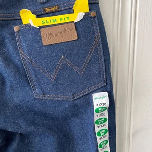 May include: Close-up of dark blue Wrangler slim fit jeans. The back pocket features a brown leather patch with the Wrangler logo. A yellow tag reads "SLIM FIT". The jeans have a size tag that reads 31x30.