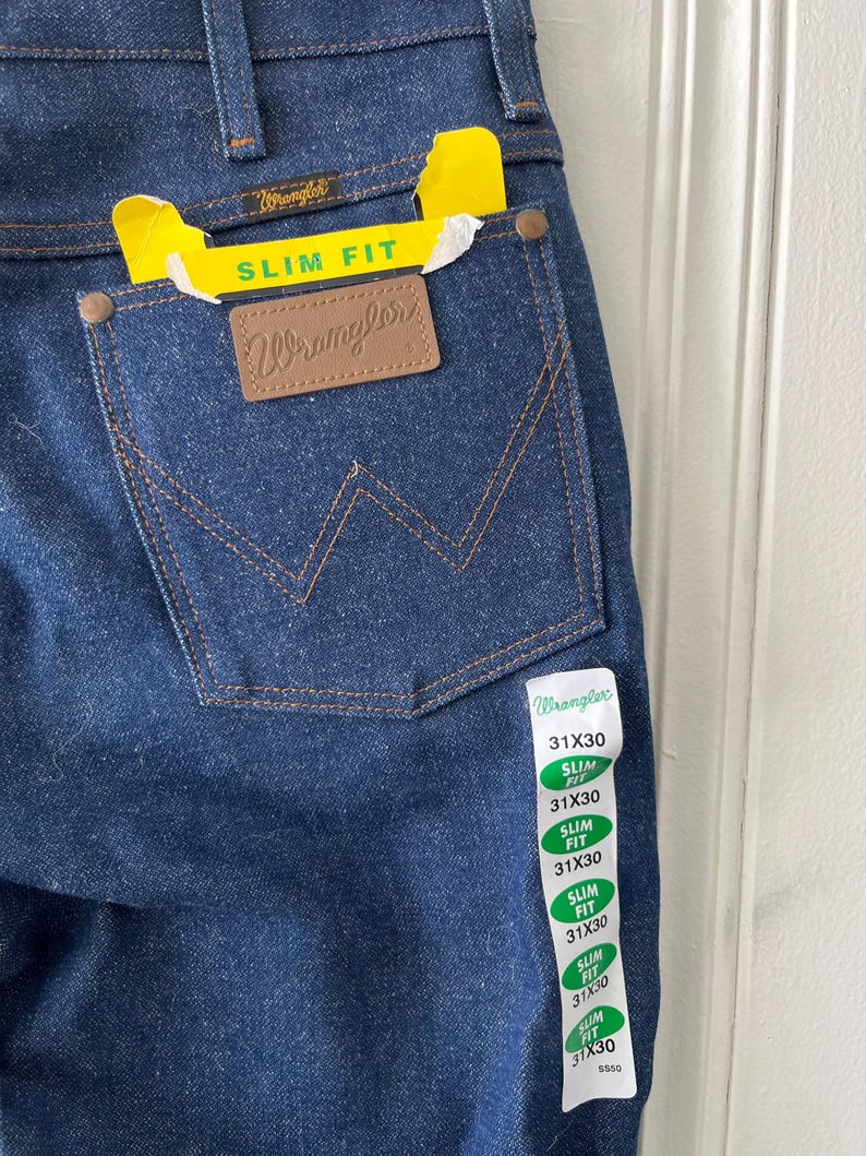 May include: Close-up of dark blue Wrangler slim fit jeans. The back pocket features a brown leather patch with the Wrangler logo. A yellow tag reads "SLIM FIT". The jeans have a size tag that reads 31x30.