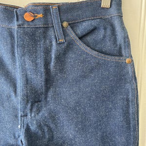 May include: Close-up of blue denim jeans. The jeans have a button closure with a copper-colored button and a small pocket on the front. The stitching is a contrasting orange color. The jeans are hanging on a metal hanger.