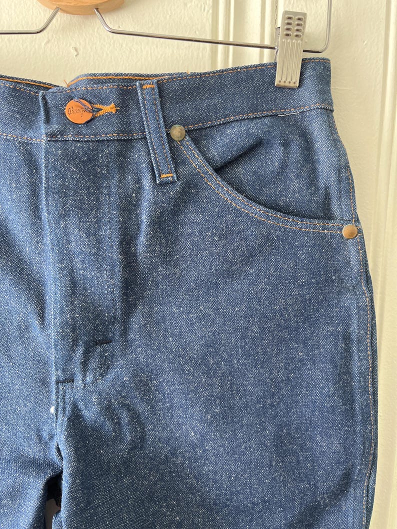 May include: Close-up of blue denim jeans. The jeans have a button closure with a copper-colored button and a small pocket on the front. The stitching is a contrasting orange color. The jeans are hanging on a metal hanger.