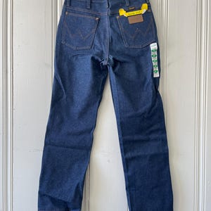 May include: Dark blue denim jeans with the Wrangler logo on the back pockets. The jeans have a straight leg fit and a yellow tag that says "SLIM FIT". The jeans are hanging on a wooden hanger.