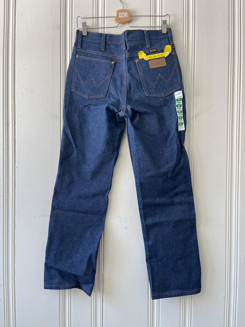 May include: Dark blue denim jeans with the Wrangler logo on the back pockets. The jeans have a straight leg fit and a yellow tag that says "SLIM FIT". The jeans are hanging on a wooden hanger.