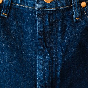 Peut inclure: Gros plan sur un jean en denim bleu fonc&eacute;. Le jean est dot&eacute; d'une fermeture boutonn&eacute;e avec un bouton de couleur cuivre et des rivets en cuivre. Les coutures sont de couleur jaune-orang&eacute; contrastante.