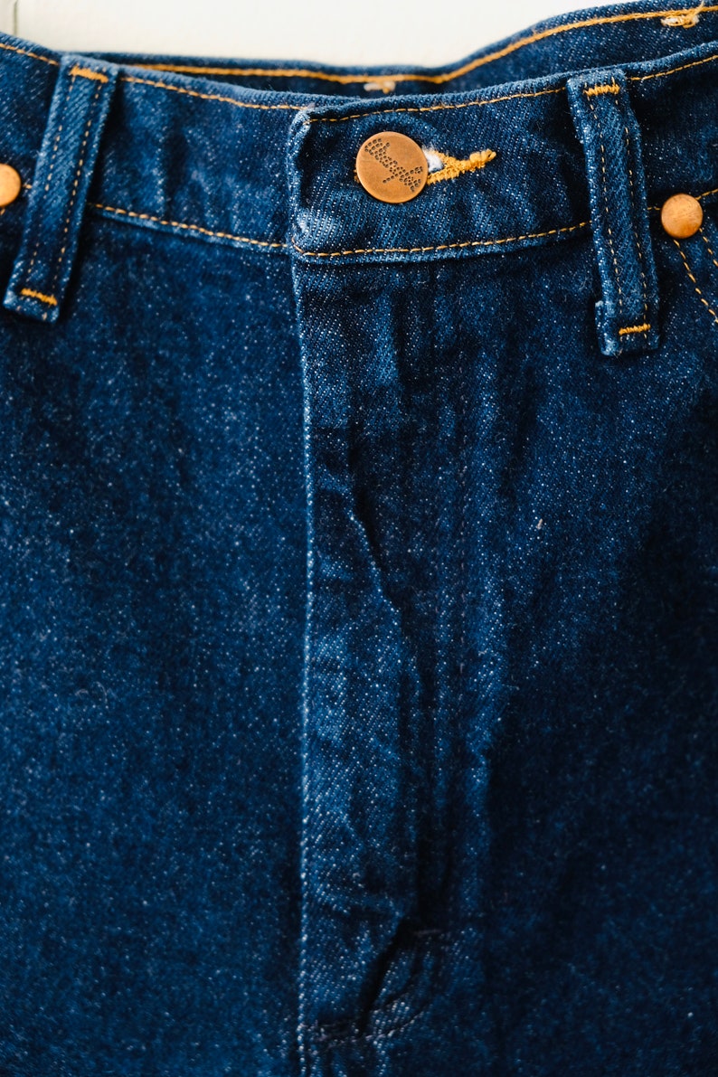 Peut inclure: Gros plan sur un jean en denim bleu fonc&eacute;. Le jean est dot&eacute; d'une fermeture boutonn&eacute;e avec un bouton de couleur cuivre et des rivets en cuivre. Les coutures sont de couleur jaune-orang&eacute; contrastante.