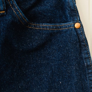 Peut inclure: Gros plan sur un tissu denim bleu fonc&eacute; avec une poche et des rivets cuivr&eacute;s. Le denim a une texture visible et est cousu avec du fil jaune. Un mat&eacute;riau classique et durable pour les v&ecirc;tements.
