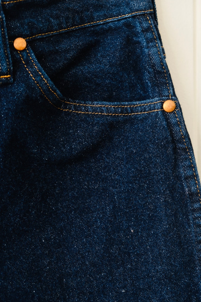 Peut inclure: Gros plan sur un tissu denim bleu fonc&eacute; avec une poche et des rivets cuivr&eacute;s. Le denim a une texture visible et est cousu avec du fil jaune. Un mat&eacute;riau classique et durable pour les v&ecirc;tements.