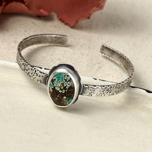 May include: A silver cuff bracelet featuring a textured, hammered finish along the band. The center of the bracelet displays an oval-shaped stone with a dark brown base and vibrant turquoise-colored inclusions, set in a smooth silver bezel.