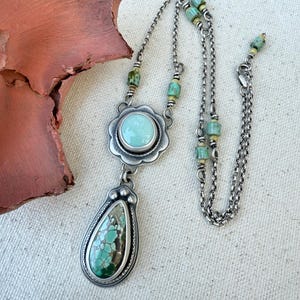 May include: A silver necklace featuring a turquoise pendant and a round turquoise stone set in a floral design. The necklace has a delicate chain with small turquoise and green beads. The pendant is teardrop-shaped with a detailed silver border.