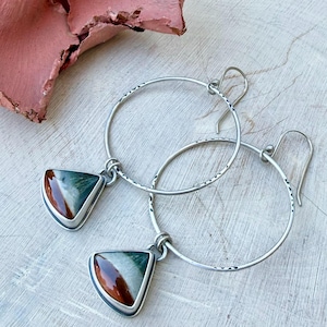 May include: A pair of silver hoop earrings with triangular pendants. The pendants feature a mix of green, brown, and white colors. The hoops have a textured design. The earrings are displayed on a light wooden surface.