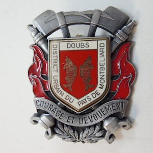 May include: A silver and red enamel badge with the text "DOUBS DISTRICT URBAIN DU PAYS DE MONTBELIARD COURAGE ET DEVOUEMENT". The badge features two crossed axes and a stylized flame design.