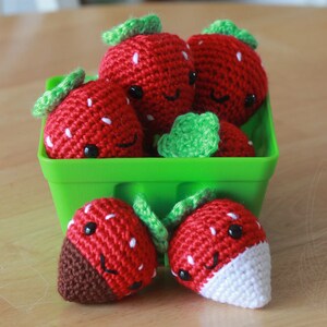 May include: A green basket filled with crocheted strawberries. The strawberries are red with white accents and have green leaves. Two strawberries are outside the basket, one with a chocolate bottom and the other with a white tip.