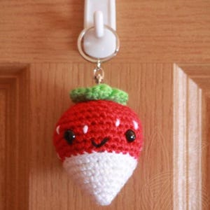 May include: A crocheted strawberry keychain with a white base and red top. The strawberry has a green stem and leaves and a smiling face with black eyes.