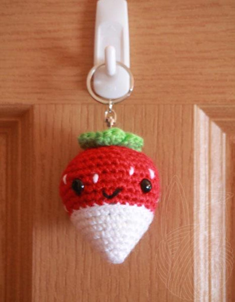 May include: A crocheted strawberry keychain with a white base and red top. The strawberry has a green stem and leaves and a smiling face with black eyes.