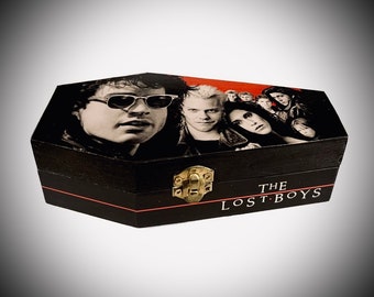 Puede incluir: Caja negra en forma de ataúd con tapa con bisagras y cierre dorado. La parte superior presenta una imagen en blanco y negro de los personajes de la película "The Lost Boys". El texto "THE LOST BOYS" está impreso en la parte delantera.