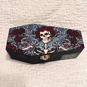 May include: Black wooden coffin-shaped box with a decorative lid featuring a skeleton with a floral crown and roses. The lid has a colorful floral pattern. The box has a gold-colored clasp and the words "Grateful Dead" on the side.