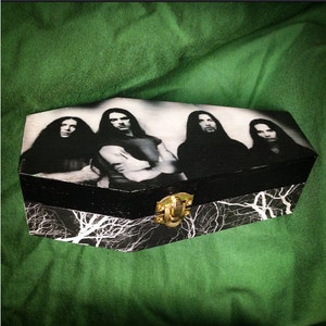 May include: A coffin-shaped box featuring a black and white band image. The box has a black top and sides, with a lightning bolt pattern on the base. It is secured with a gold-coloured clasp.