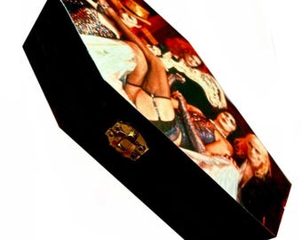 May include: A black coffin-shaped wooden box featuring a hinged lid with a gold-tone metal latch. The top surface displays a colorful, detailed photographic print of figures in elaborate costumes, including individuals with red hair and dark makeup.