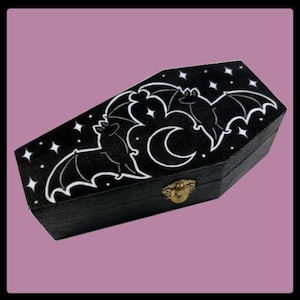 May include: Black coffin-shaped box with a bat and moon design. The top of the box features a white illustration of two bats, a crescent moon, and stars. The box has a gold-colored clasp and is set against a pink background.
