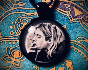 May include: Black pendant necklace featuring a black and white image of a person. The round pendant is set in a black metal frame and hangs from a black cord. The background is a patterned blue and gold fabric.