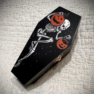 May include: Black coffin-shaped box with a Halloween theme. The box features a white skeleton holding two orange jack-o'-lanterns. The coffin has a gold-coloured clasp. The design is on a black background.