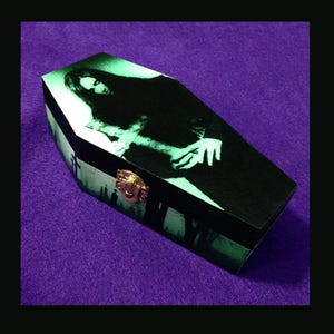 May include: A black wooden coffin-shaped box with a hinged lid and a gold-coloured clasp. The lid features a green-tinted image of a figure with outstretched hands. The sides of the box have a green silhouette design.