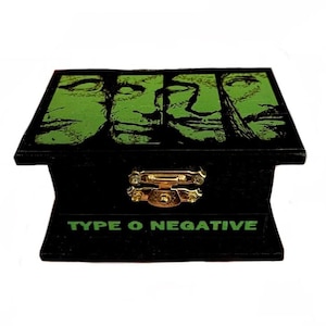 Può includere: Scatola di legno nera con coperchio incernierato. Il coperchio presenta una grafica verde e nera di volti. La scatola ha una chiusura color oro e le parole "TYPE O NEGATIVE" in lettere verdi sul davanti.