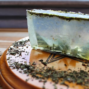 May include: A clear, rectangular bar of soap with green flecks and gold glitter. The soap is partially cut, revealing a layered interior. The soap rests on a wooden surface with scattered dried herbs.