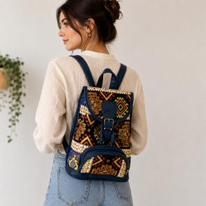 May include: A backpack featuring a dark blue faux leather trim and a woven fabric panel with a multi-colored geometric pattern in shades of black, gold, and white. The bag includes a top flap with a buckle closure and a front zippered pocket.