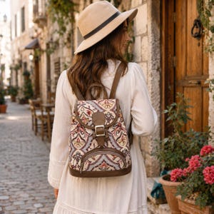 May include: A beige fedora hat with a black band and a backpack featuring a multi-colored floral tapestry pattern, brown leather-like trim, a top flap with a metal buckle closure, and a front zippered pocket, worn over a long-sleeved white dress.