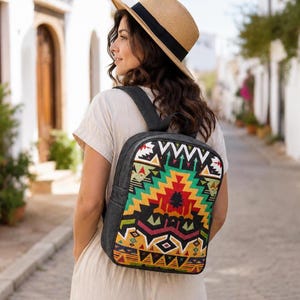 Puede incluir: Una mochila con un vibrante patrón geométrico multicolor en tonos amarillo, verde, rojo y negro en el panel posterior, con paneles laterales y correas de hombro en negro sólido, sobre una prenda de color beige claro y un sombrero de paja.