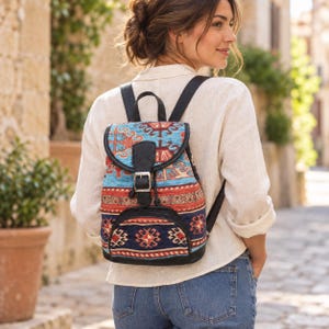 Könnte beinhalten: Ein kleiner Rucksack aus gewebtem Stoff mit geometrischen Mustern in Blau-, Rot- und Cremetönen. Die Tasche verfügt über schwarze Kunstlederbesätze, eine Klappe mit silberfarbenem Metallschnallenverschluss und verstellbare schwarze Schultergurte.