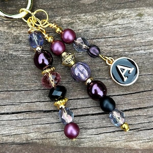 May include: A gold-toned keychain with three beaded strands. The strands feature a mix of purple, black, and clear beads, accented with gold-colored spacers. One strand has a charm with the letter "A" on a black background.