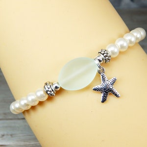 May include: A beaded bracelet featuring white faux pearls and a central, pale green, oval-shaped bead. A silver-colored starfish charm hangs from the center. The bracelet has silver-colored decorative beads.