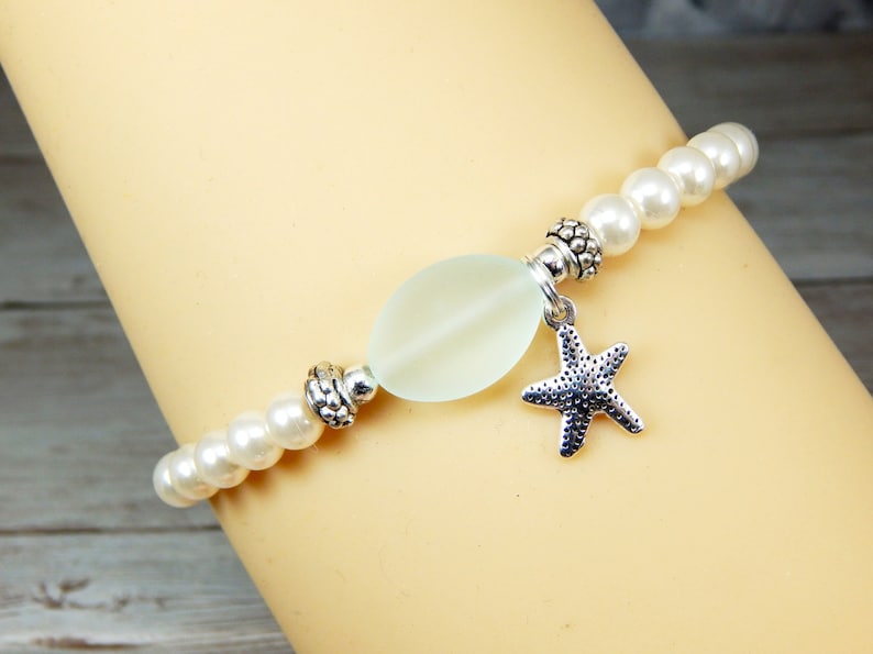 May include: A beaded bracelet featuring white faux pearls and a central, pale green, oval-shaped bead. A silver-colored starfish charm hangs from the center. The bracelet has silver-colored decorative beads.