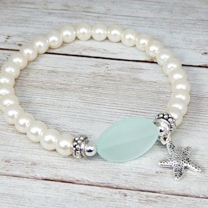 May include: A white pearl bracelet with a light blue, oval-shaped bead and a silver starfish charm. The bracelet is strung on an elastic band. The bracelet is on a light wood surface.