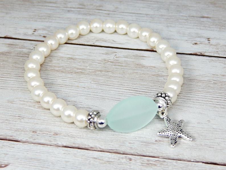 May include: A white pearl bracelet with a light blue, oval-shaped bead and a silver starfish charm. The bracelet is strung on an elastic band. The bracelet is on a light wood surface.