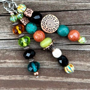 May include: A collection of beaded keychains with various colorful beads, including green, orange, turquoise, and black. The keychains feature a mix of shapes and textures, with metal accents and a spiral design.