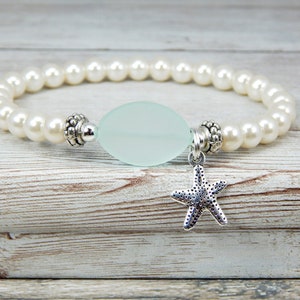 May include: A beaded bracelet featuring faux pearls and a pale green oval-shaped bead. A silver starfish charm hangs from the center. The bracelet is displayed on a light wood-grain surface.