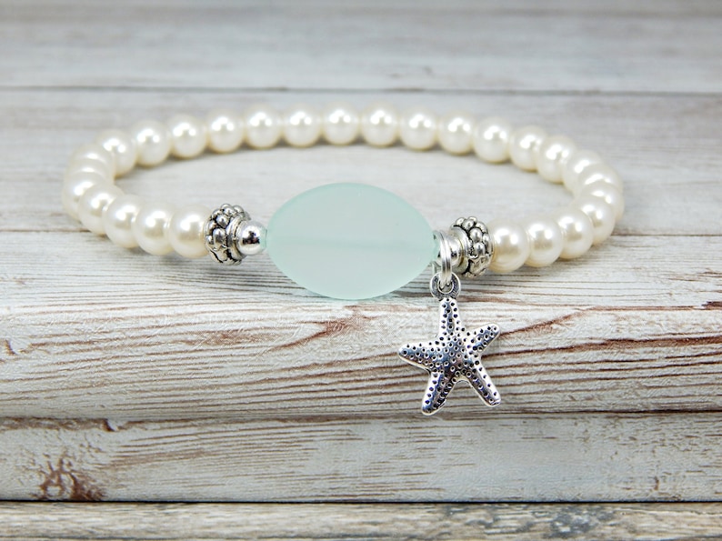 May include: A beaded bracelet featuring faux pearls and a pale green oval-shaped bead. A silver starfish charm hangs from the center. The bracelet is displayed on a light wood-grain surface.