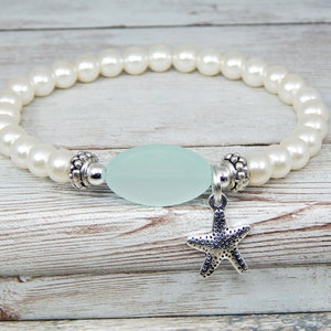 May include: A stretch bracelet featuring white faux pearls, a light blue oval bead, and a silver starfish charm. The bracelet is designed with a beach or nautical theme, perfect for everyday wear or special occasions.