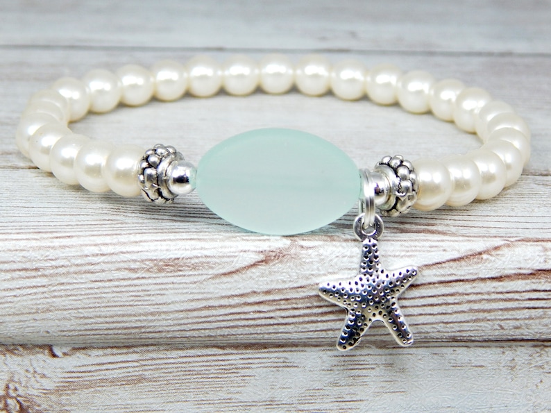 May include: A stretch bracelet featuring white faux pearls, a light blue oval-shaped bead, and a silver starfish charm. The bracelet is designed with silver accents and is suitable for everyday wear.