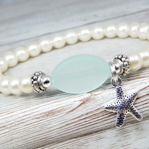 May include: A beaded bracelet featuring creamy white faux pearls. The bracelet has a pale blue, oval-shaped bead in the center, flanked by decorative silver-toned metal accents. A small, silver starfish charm dangles from the bracelet.