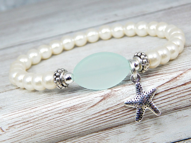 May include: A beaded bracelet featuring creamy white faux pearls. The bracelet has a pale blue, oval-shaped bead in the center, flanked by decorative silver-toned metal accents. A small, silver starfish charm dangles from the bracelet.