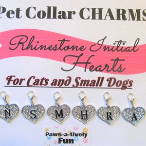 May include: Six silver heart-shaped charms with rhinestones and a black letter on each charm. The letters spell out "NSMHRA". The charms are for cats and small dogs. The charms are on a white background with a pink banner that says "Rhinestone Initial Hearts". Below the charms is a small white tag that says "Paws-a-tively Fun".