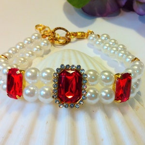 Könnte beinhalten: Ein doppelreihiges Perlenarmband mit einem großen roten Strassstein in einem goldenen Rahmen und zwei kleineren roten Strasssteinen auf jeder Seite.