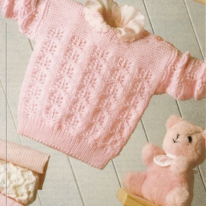 May include: A pink knitted baby sweater with a delicate lace pattern. The sweater has a white lace collar and is hanging on a wooden hanger.